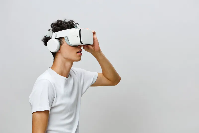 Молодой человек, погружённый в опыт виртуальной реальности, в белом VR-шлеме и наушниках на фоне минималистичного серого фона, что демонстрирует современные технологии и концепции иммерсивных игр