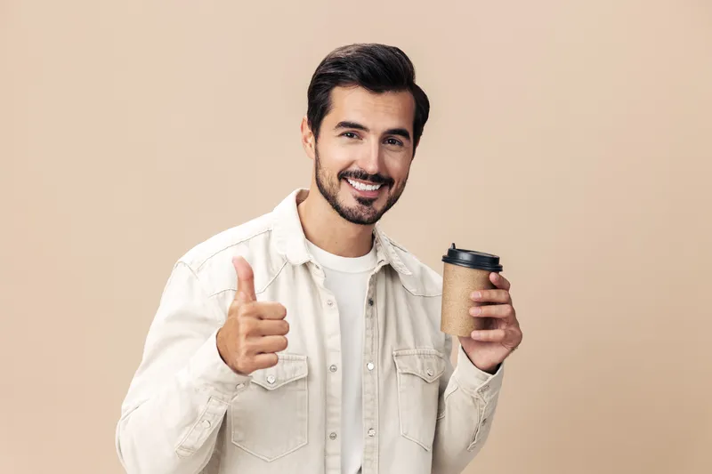 Портрет стильного мужчины в белой футболке, который держит стакан с кофе, на бежевом фоне