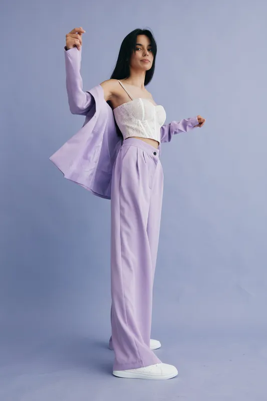 Young woman wearing stylish lilac pants and blazer with a white lace top posing confidently against a soft blue background in a modern fashion shoot.