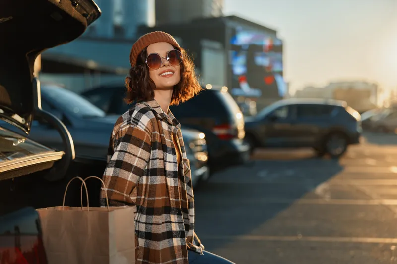 Woman shopping at car in parking lot with bags, smile and sunlight; candid lifestyle portrait with authenticity, mindful living and golden hour glow capturing emotional storytelling.