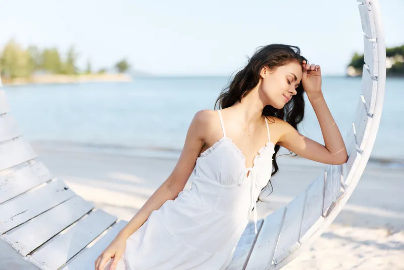 [A graceful woman in a white dress lounges on a curved wooden frame at the beach, sunlight highlights the soft fabric and relaxed pose against a tranquil shoreline.]