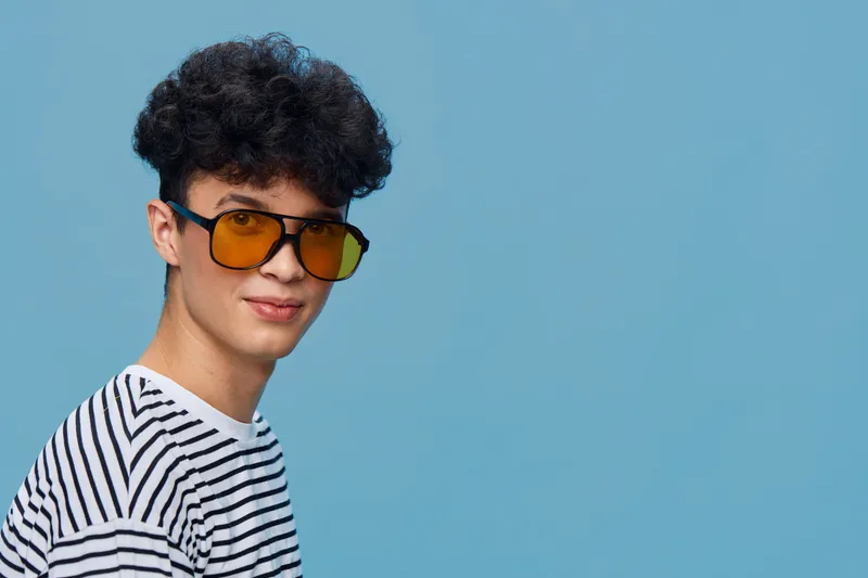 Young man wearing yellow sunglasses and striped shirt stands against blue background with curly hair and casual expression, modern style portrait.