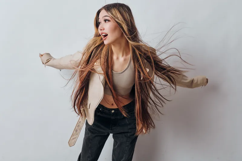 Young woman with long hair in casual jacket and black pants dancing or posing with energetic and joyful expression on light background in studio environment with flying hair.