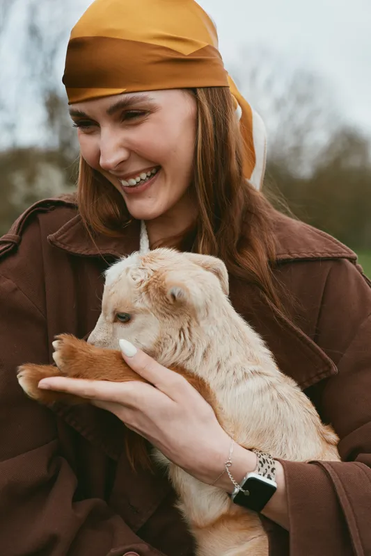 A joyful woman in a warm headscarf holds a young goat kid outdoors, smiling as they share a tender moment. A candid lifestyle scene of animal bond and care.