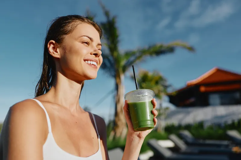 Smiling woman outdoors drinks a green smoothie, enjoying sunshine on a bright garden setting, wellness and lifestyle in focus, hydration and healthy living tone, natural light creating a vibrant