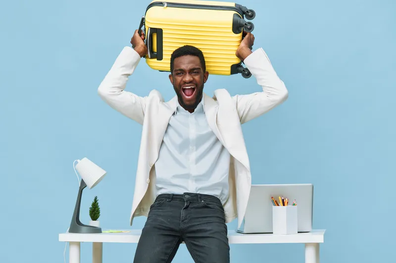 Happy young African American man holding yellow suitcase above his head, excited expression, standing in modern office space with desk, laptop, and stationery, bright mood, casual business style. Peop