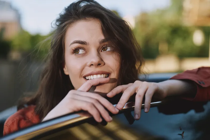 A young woman leans on a car window outdoors, smiling softly as warm sunlight highlights her features and casual style in a candid lifestyle moment