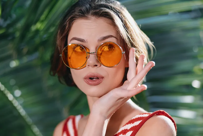 A cheerful woman wearing round orange sunglasses poses outdoors among tropical leaves, smiling warmly and exuding a playful, sunny style and relaxed confidence.
