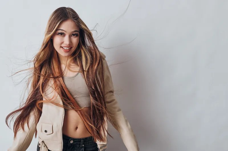 Young woman with long brown hair smiling and posing in casual beige jacket and cropped top with grey background for fashion portrait and style.