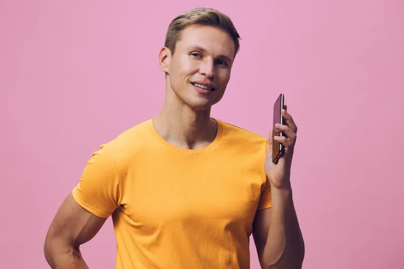 Cheerful smiling young man with short light brown hair, wearing bright yellow t-shirt, holding smartphone in hand, looking at camera, posing against pastel pink background, studio portrait, casual sty