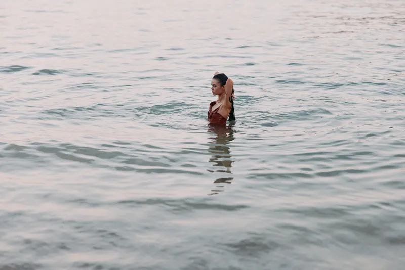 A lone woman stands waist deep in clear sea water, soft light glinting on the surface as gentle waves cradle her silhouette, creating a serene, intimate moment.