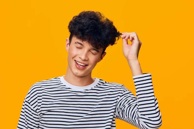 young man with curly hair wearing striped shirt smiling joyfully on vibrant orange background expressing happiness and carefree mood in casual outfit