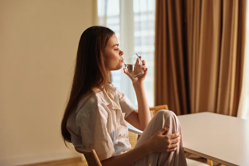 Woman sitting at home, relaxed portrait drinking water from a glass by a window, natural sunlight, casual outfit, peaceful morning moment and calm domestic lifestyle.
