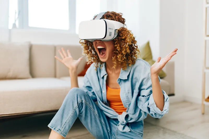 Улыбающаяся женщина наслаждается игрой в виртуальной реальности дома, сидя на диване в VR-очках и с контроллером в руках