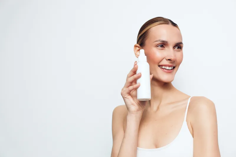 Smiling woman holding white cosmetic bottle with dropper applies skincare product on face isolated on white background with natural makeup and clean fresh skin.