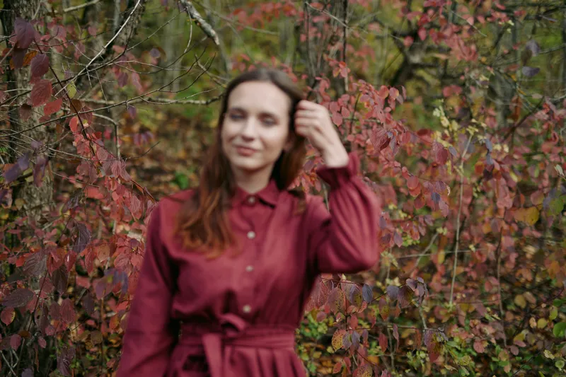 A candid natural portrait of a woman in a red dress standing among autumn foliage, conveying genuine presence and unposed character in a soft, intimate outdoor moment of truth.