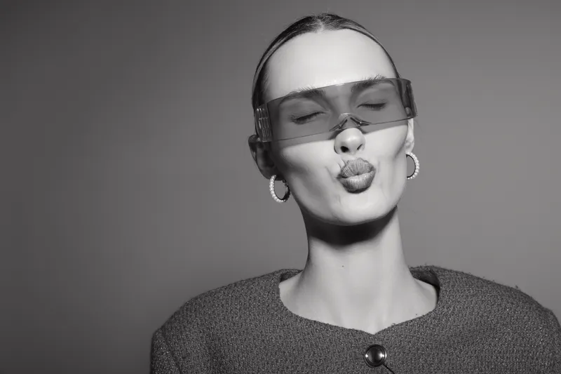 Young white woman with pale skin wearing large sunglasses and hoop earrings makes a playful kiss face on grey background.