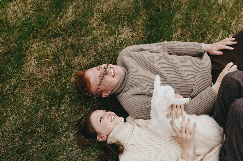 A couple lounges on grass outdoors with a small goat kid cuddled between them, smiling and relaxed. A cozy lifestyle moment showing gentle animal companionship and simple joy in nature.