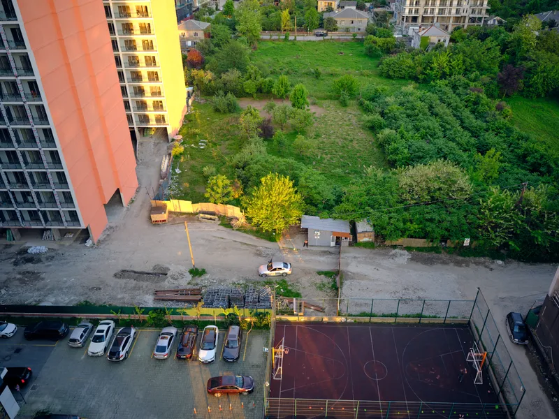 Building under construction by parking lot with cars and fenced court beside green vacant lot, road and small shed seen from aerial urban view at golden hour, modern city scene