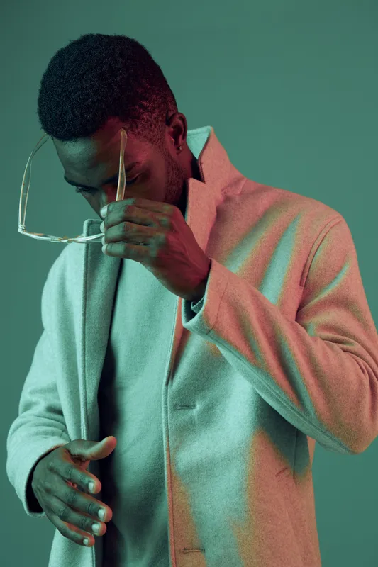 Stylish African man in a grey coat, adjusting stylish glasses against a teal background, conveying a sense of confidence and modern fashion ideals
