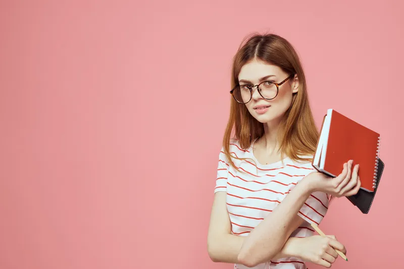 Красивая студентка держит книги, жестикулирует руками на розовом фоне