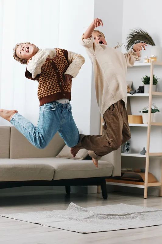 Two children leap midair in a bright living room, laughter filling the space as they bounce on a sofa in cozy knit sweaters and jeans, capturing carefree childhood energy.