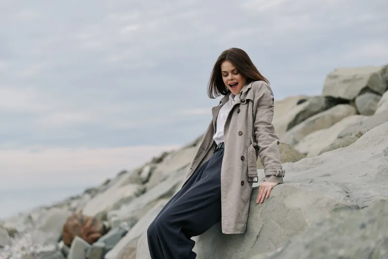 Young woman in casual clothes sits on large rocks outdoors under cloudy sky, looking down with a surprised expression. Concept of nature, fashion, and outdoor lifestyle.