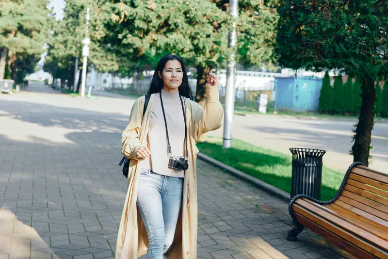 A woman in a long beige coat and light denim jeans walks along a sunlit park path with a camera hanging, showcasing casual outdoor urban fashion and a relaxed lifestyle moment