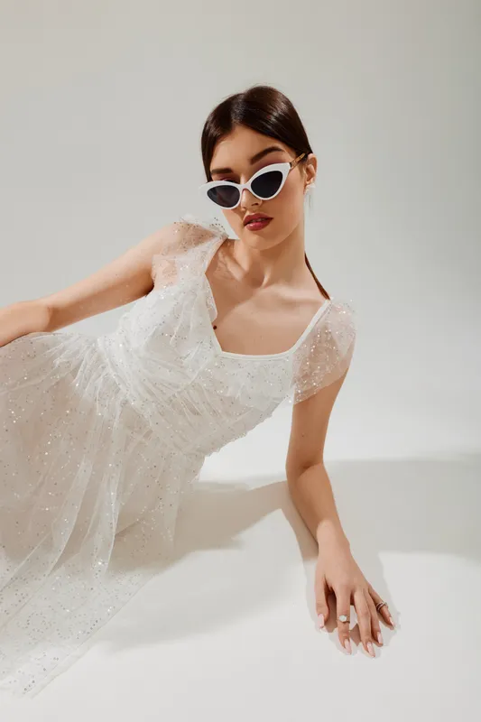 Young woman in a white sparkling dress and stylish sunglasses poses on a white background. Concept of woman beauty fashion, elegance, and modern style in studio lighting.