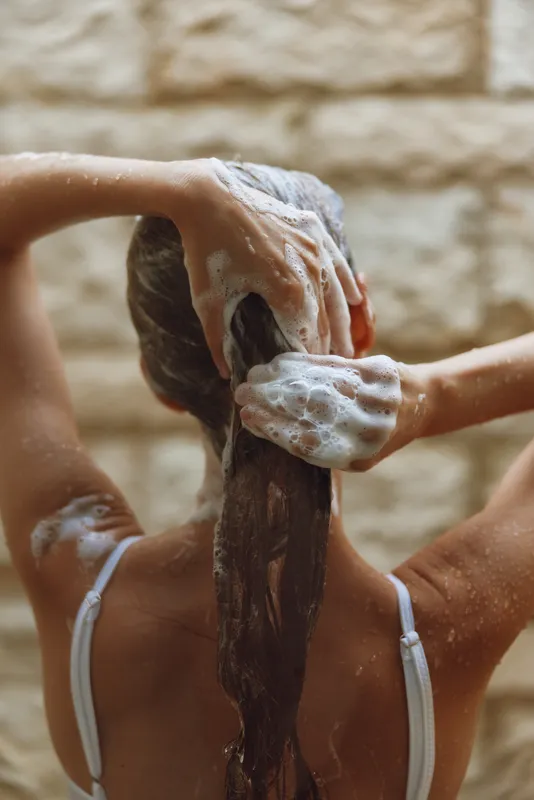 Woman washing long hair with shampoo foam, hands massaging scalp, wet strands, natural light, indoor bathroom with textured wall background.