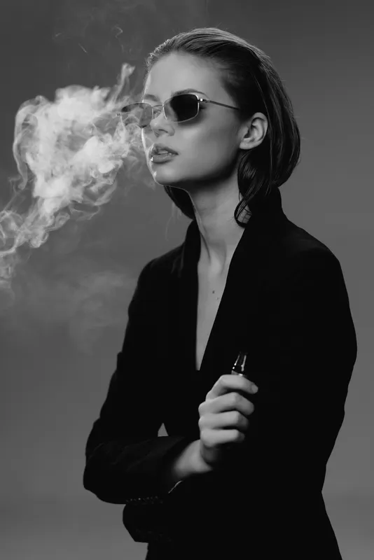 Black and white portrait of a woman vaping with sunglasses in a dark outfit, exhaling smoke in a studio setting.