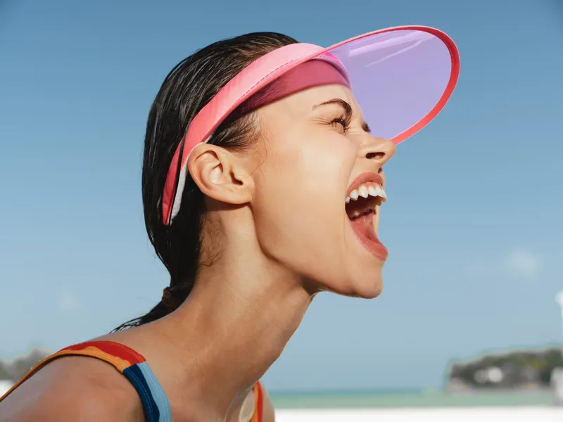 Close up portrait of a joyful woman at the beach, wearing a pink visor, mouth wide open in laughter, sunlit skin, damp hair, and a bright blue sky behind, energetic mood.