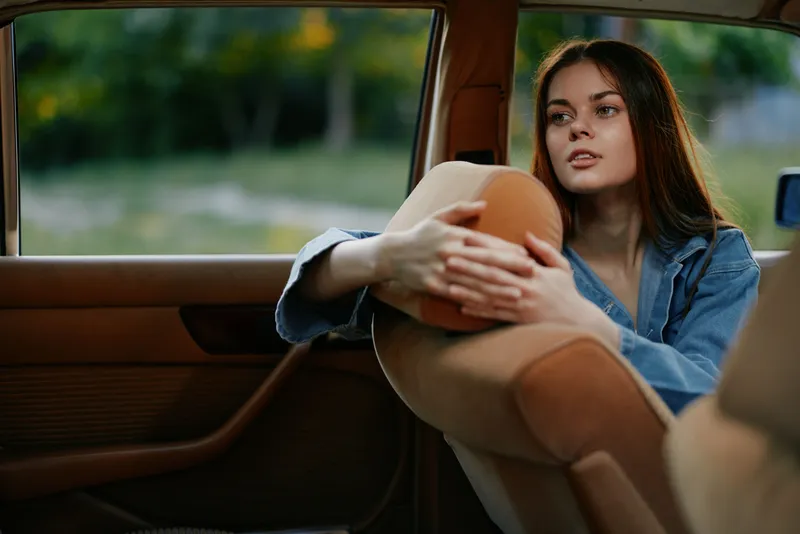 Thoughtful young woman in a denim jacket looking out of a vintage car window, showcasing emotions of reflection and nostalgia in a serene outdoor setting.
