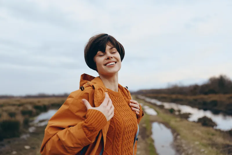 Woman outdoors portrait, smiling in orange jacket and scarf, walking beside a river path, soft daylight countryside lifestyle moment, travel nature background