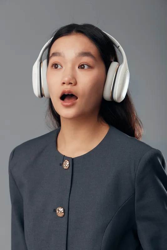 Young woman wearing headphones with surprised expression in casual dark jacket against gray background. Portrait of girl with long hair listening to music or audio content.