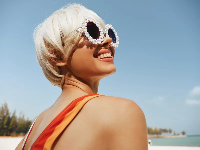 Carefree beach scene focusing on a smiling woman with short blonde hair and glittery heart sunglasses, wearing an orange swimsuit, set against a bright blue sky and warm sunlight.