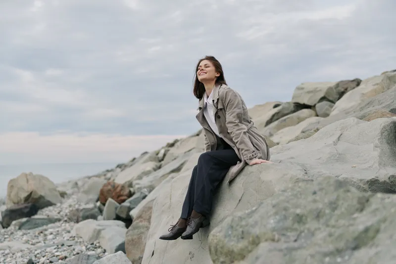 Young woman in casual jacket sits on large rocks near pebble beach under cloudy sky. She smiles and looks relaxed, enjoying outdoor nature and peaceful coastal atmosphere.
