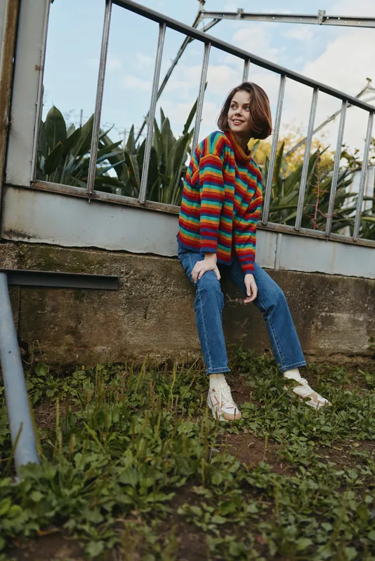 Woman colorful sweater jeans sitting smile outdoor fence casual girl portrait full length short hair sandals relaxed in garden on wall, smiling and looking away in natural daylight.