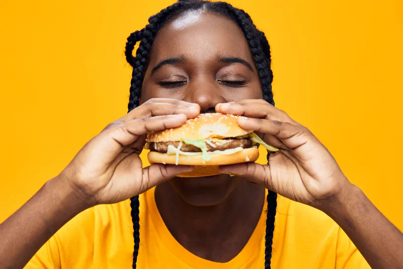 Happy young Black female enjoying a delicious burger against a vibrant yellow background, expressing joy with closed eyes, showcasing healthy eating concepts and vibrant colors