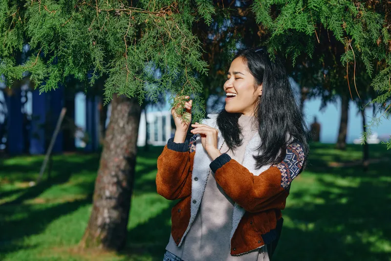 Woman outdoors near a tree, smiling and touching leaves, wearing a casual jacket, bathed in sunlight, capturing nature warmth and a candid park moment
