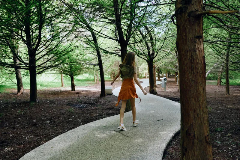 Woman walking path forest backpack dress trees trail nature outdoors summer sneakers solitude wander exploration with serene sunlight on curved pathway for relaxed outdoor journey.