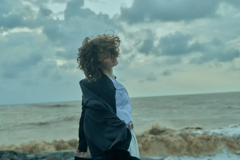 Young woman in a dark blazer and white shirt stands on a windy coastline, hair swept by sea breeze, exploring fashion and travel mood by the ocean.
