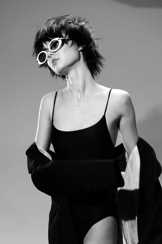 Black and white portrait of a stylish young woman wearing sunglasses and a black dress with a jacket draped over her shoulders.