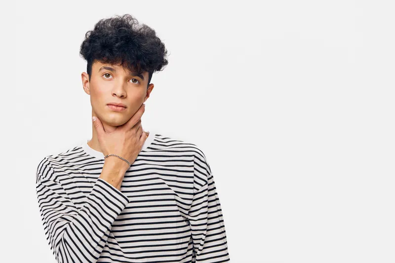 young man in striped shirt touching neck with thoughtful expression, curly hair, minimalistic white background, casual style, natural lighting