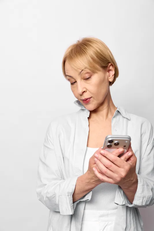 Blonde elderly woman with smartphone in portrait, indoors on white background, looking down with concerned expression, holding phone with both hands, soft even lighting