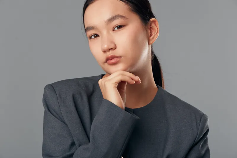 Young woman in business attire with thoughtful expression and hand near face posing against gray background for professional portrait and confident look in studio.