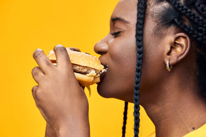 Young woman with braids delightfully enjoying a delicious hamburger against a vibrant yellow background, showcasing joy and culinary indulgence