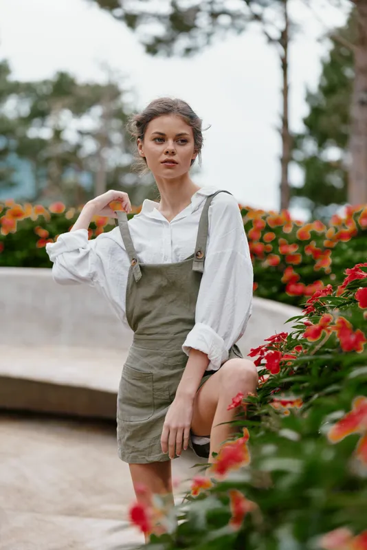 Young woman in a park setting, wearing a white shirt and green overalls, surrounded by colorful flowers, expressing a thoughtful emotion, evoking a sense of nature