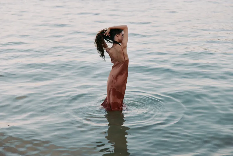 Graceful figure stands waist deep in tranquil water, wearing a flowing gown. Back view showcases wet fabric clinging to skin, soft ripples, and a dreamy, contemplative mood.