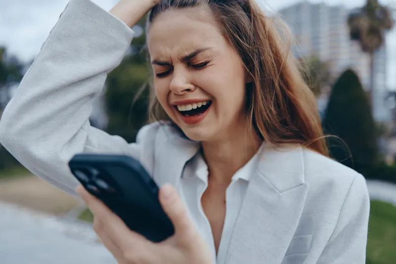 Young woman in light blazer expressing frustration and stress while looking at smartphone screen outdoors with city background, emotional business crisis concept